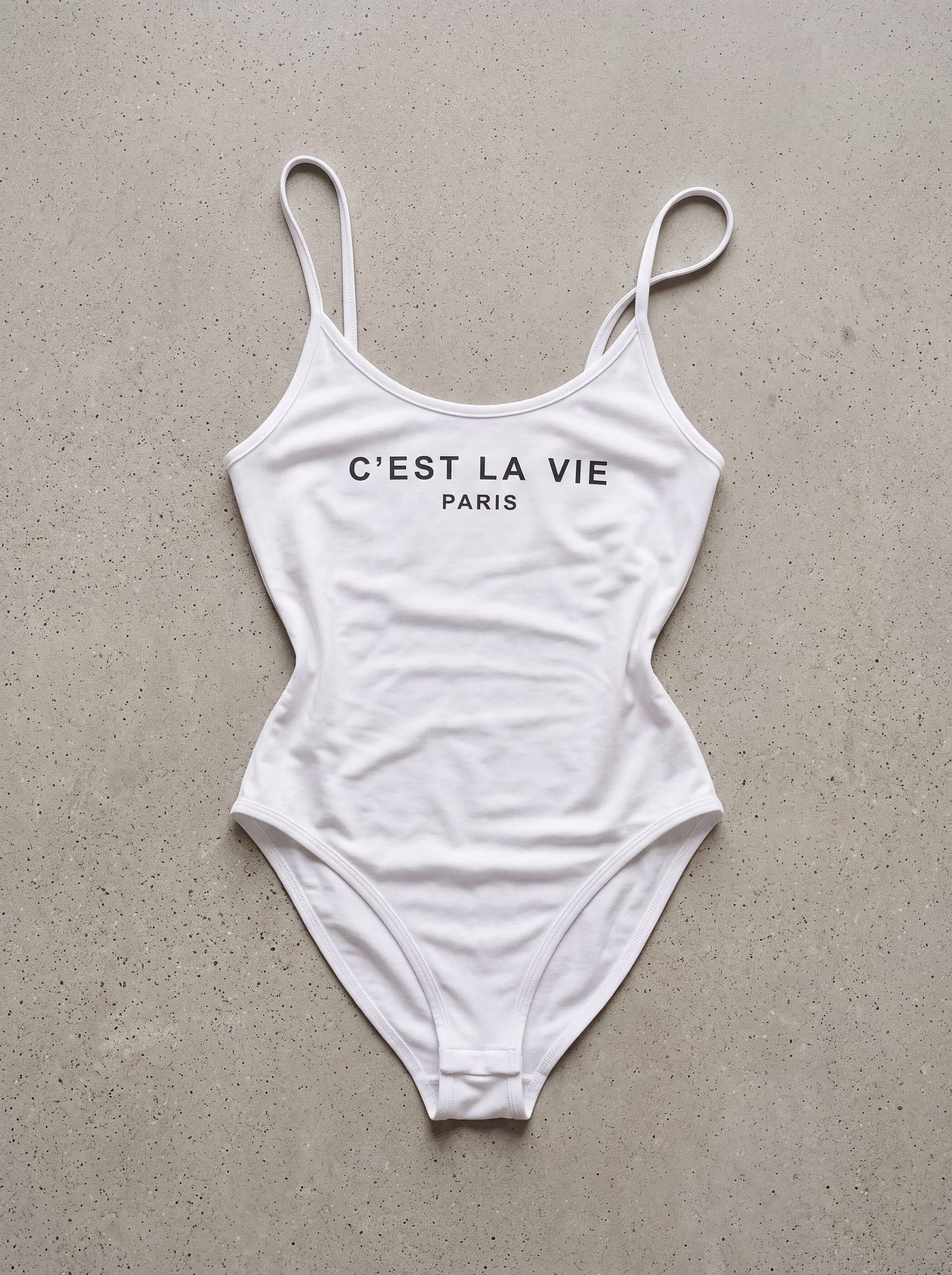 Body tirantes cuello cest la vie, en color blanco, prenda sola sobre fondo de cemento