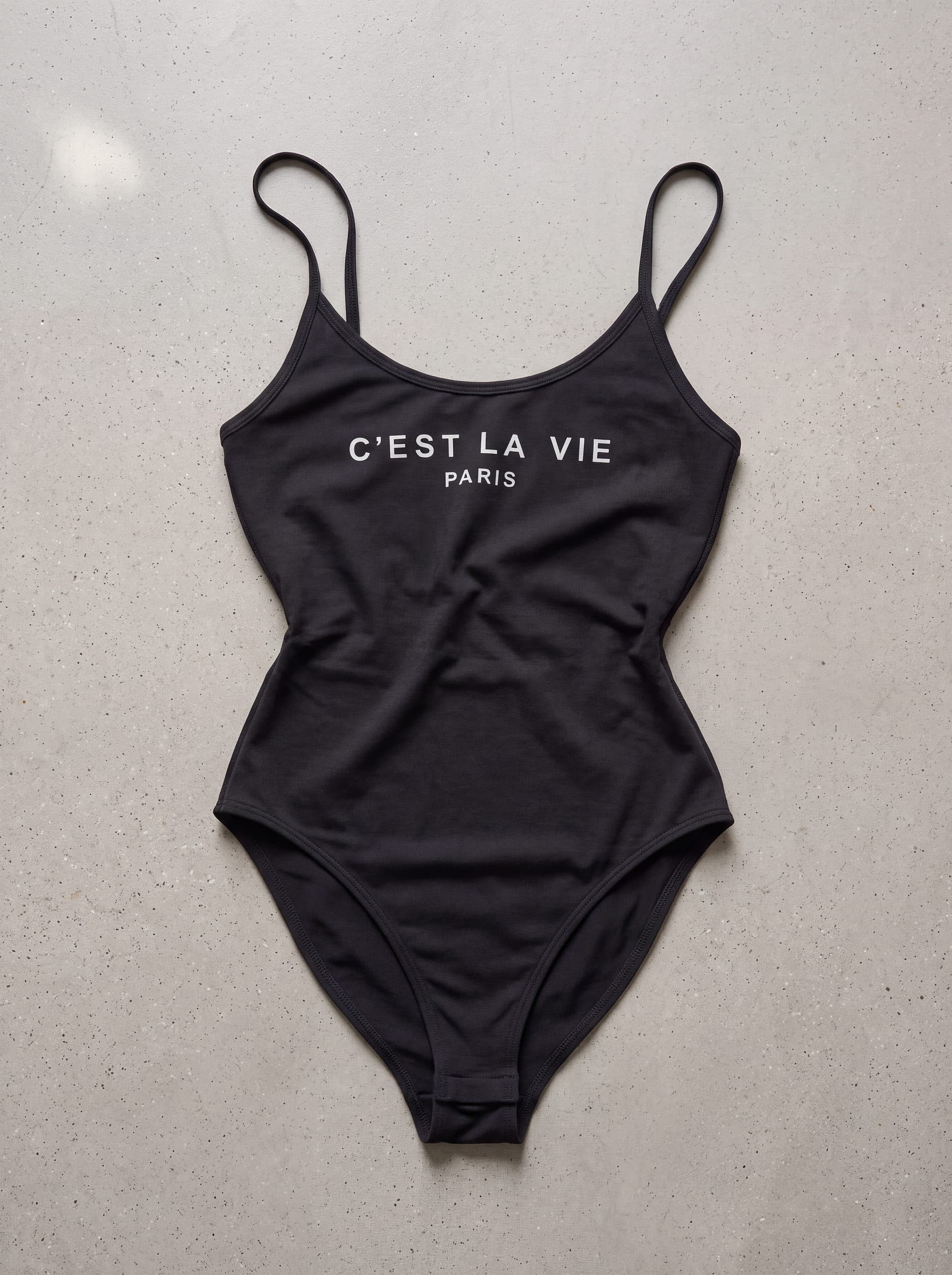 Body tirantes cuello cest la vie, en color negro, prenda sola sobre fondo de cemento
