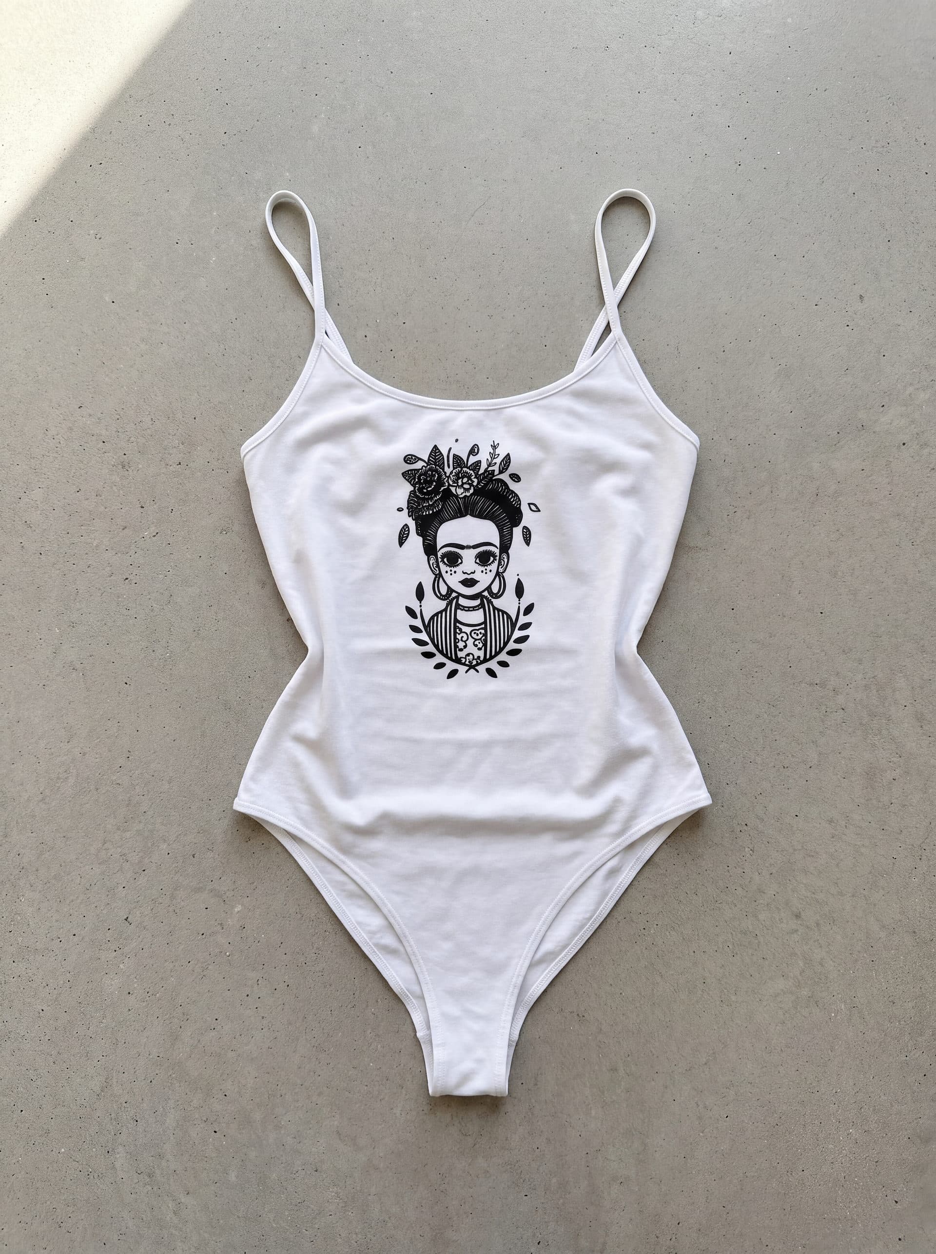 Body tirantes Frida Kahlo, en color blanco, prenda sola sobre fondo de cemento