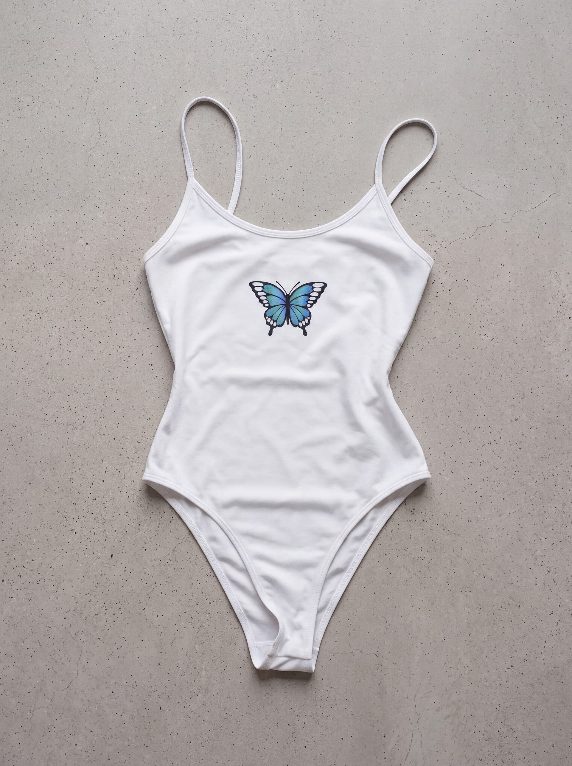 Body tirantes Mariposa Azul en el centro, en color blanco, prenda sola sobre fondo de cemento