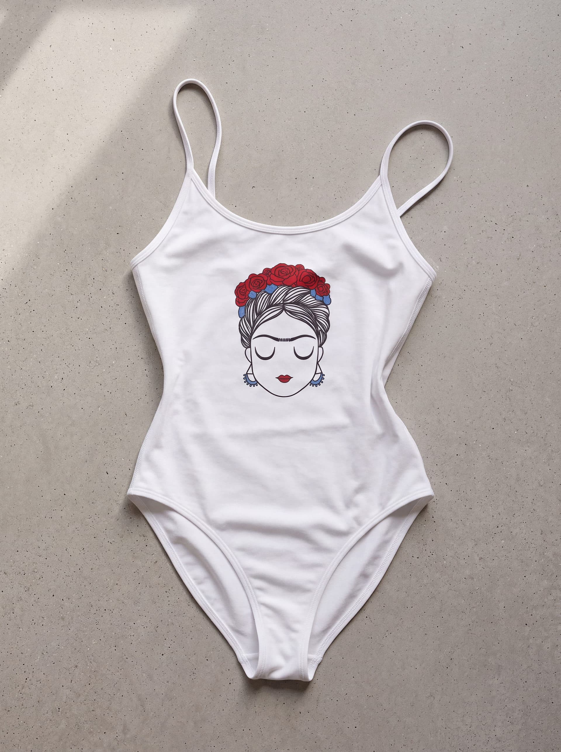Body tirantes Frida Kahlo rosas, en color blanco, prenda sola sobre fondo de cemento